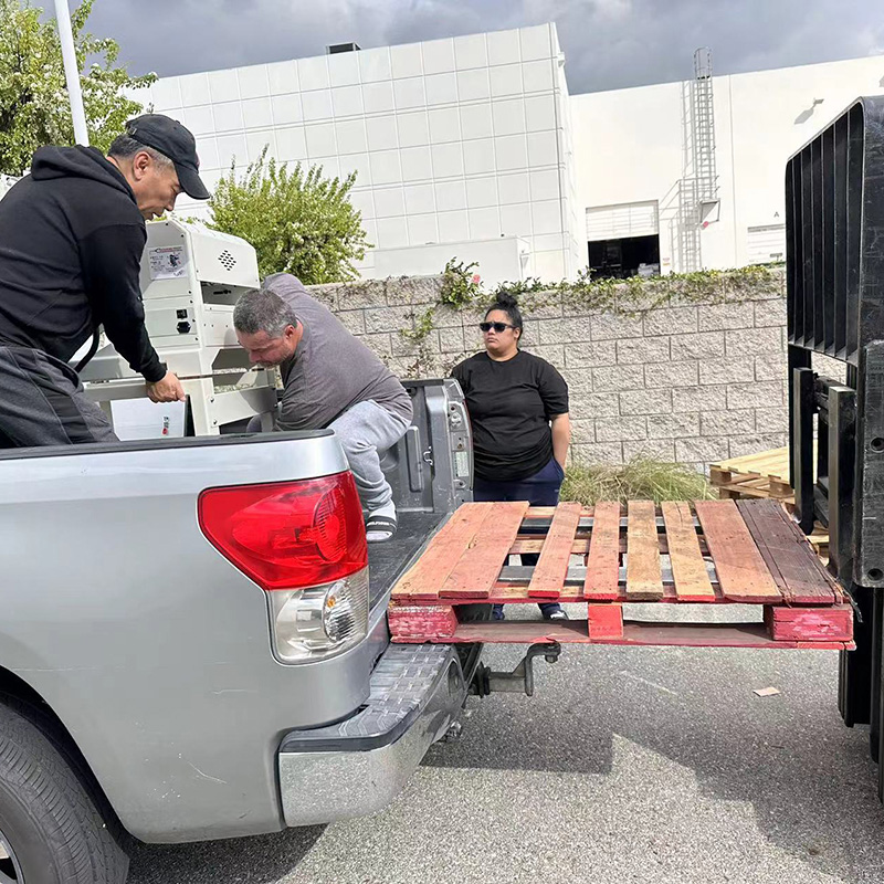 Three people loading items onto a wooden pallet in the bed of a silver pickup truck, with a building and trees in the background